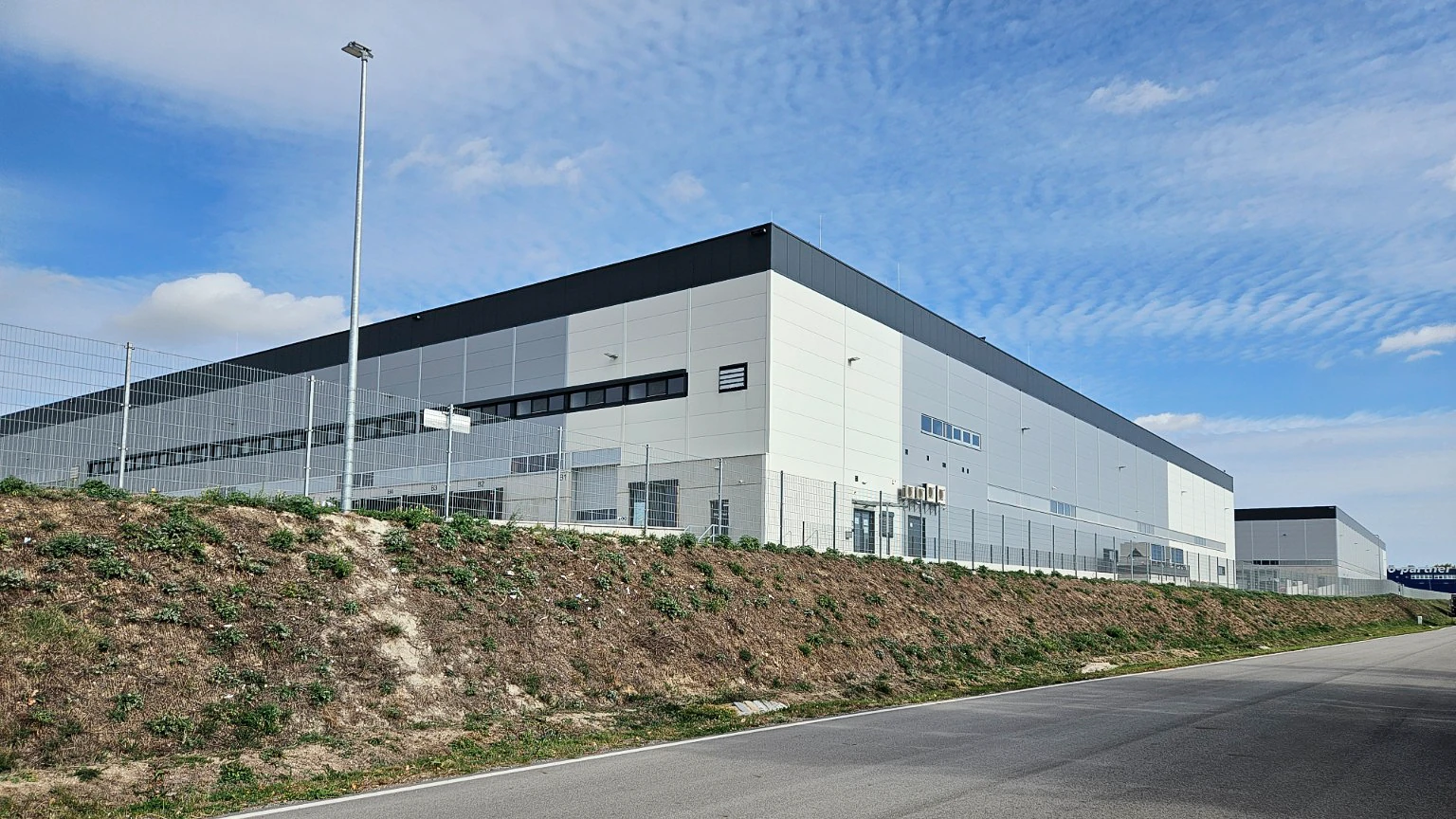 Light-grey logistics warehouse with windows, perimeter fence and an access road.