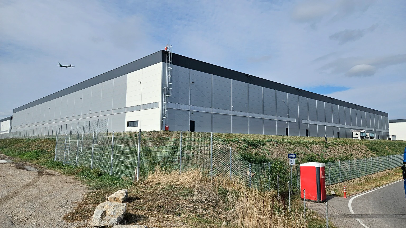 Two facades of a logistics warehouse behind a fence, with an aircraft visible in the sky.