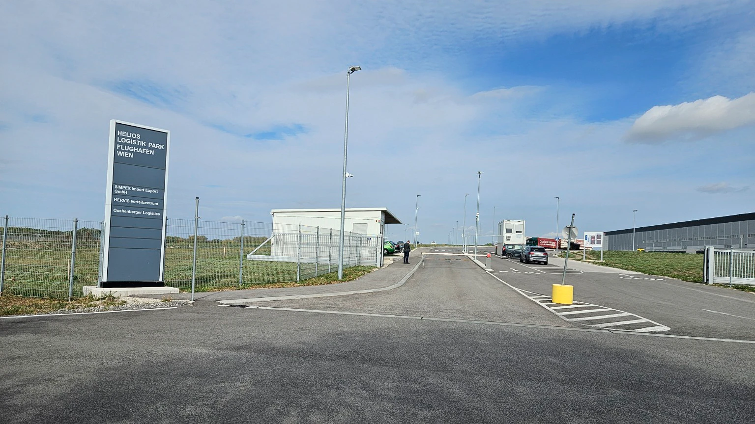 Entrance to a logistics site with a directory sign, gatehouse and warehouse buildings.